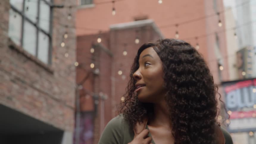A woman wearing jeans and gold sneakers walks down an urban alleyway then turns to look around with a brown backpack over her shoulder and string lights overhead