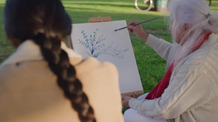 An elderly woman sits in a sunlit field at midday carefully sketching a plant with branches and leaves onto a canvas beside her a young woman watches attentively