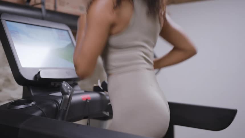 A young woman runs on a treadmill in the gym at midday her movements steady and focused as the bright light filters in highlighting the energy of her workout