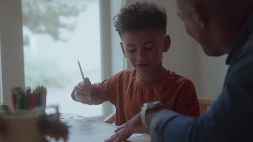 Child draws or writes with colored pencils as adult attentively guides in sunlit setting