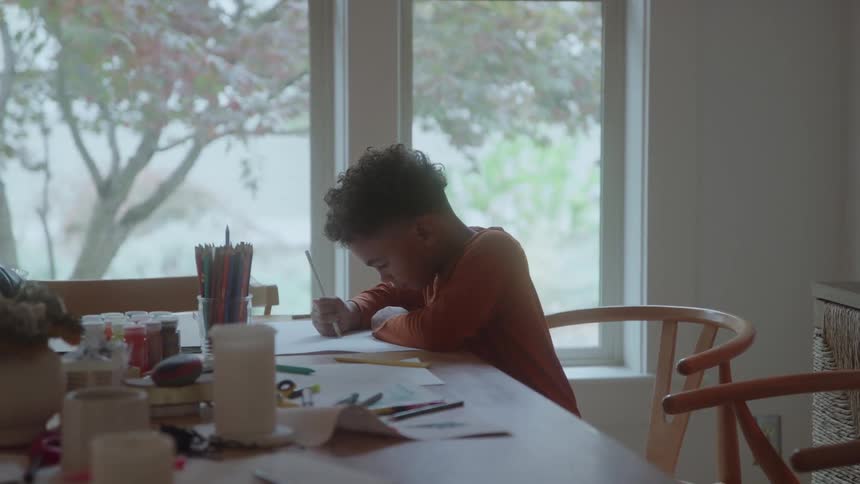 A kid picks a pen from a bowl filled with pens on the dining table and begins writing on a piece of paper focused on the task at hand