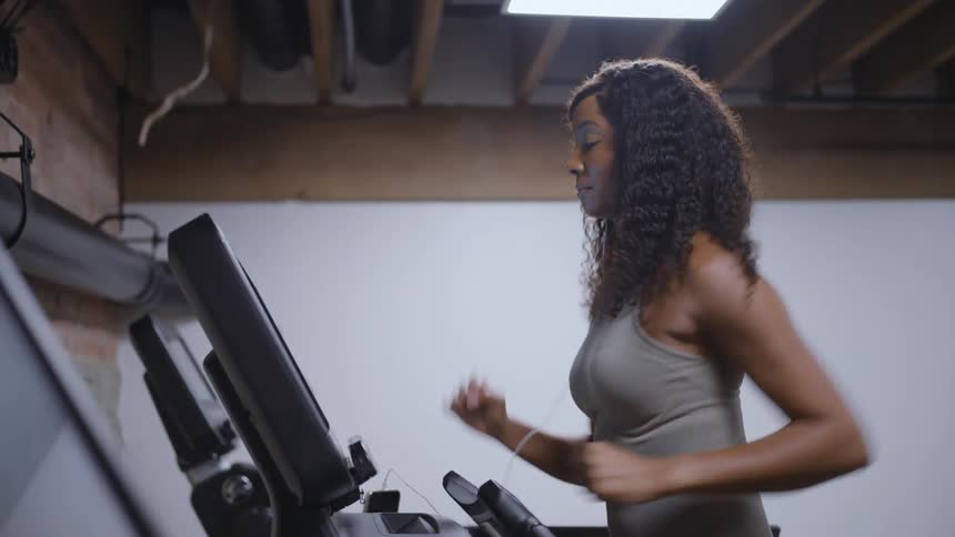 A young black woman with long curly hair runs on a treadmill in a gym wearing an ashcolored singlet and white wired earbuds her stride is focused and energetic as she moves to the rhythm of her workout