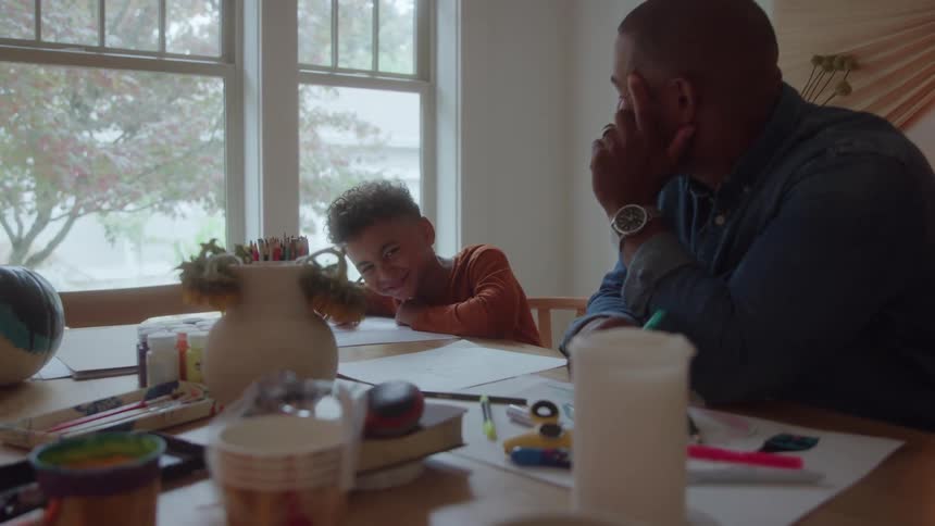 A father shares a joyful moment with his son as they sit together at the dining table both leaning over sheets of paper and writing