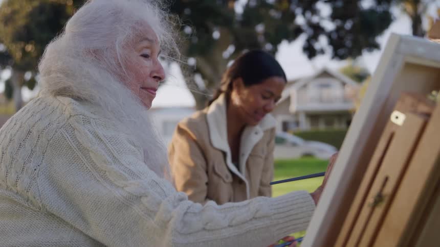 Elderly woman painting next to a younger adult woman at a park outdoors during sunset