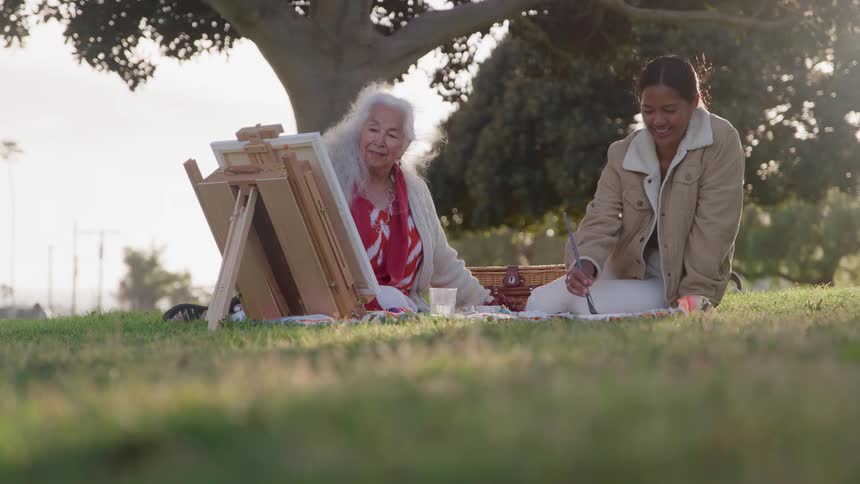 An older woman with long white hair paints on a small easel while a younger woman sits next to her smiling on a blanket on the grass beneath a large tree during golden hour