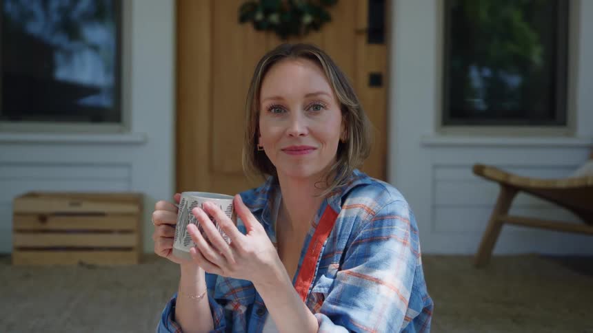 Woman holding a decorated mug with both hands sitting in front of a door with a wreath