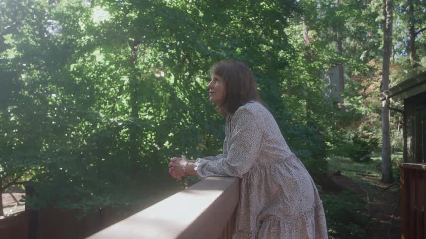 A woman in a flowing white and black patterned dress leans on a railing outdoors looking up and smiling softly with bright sunlight shining through green trees