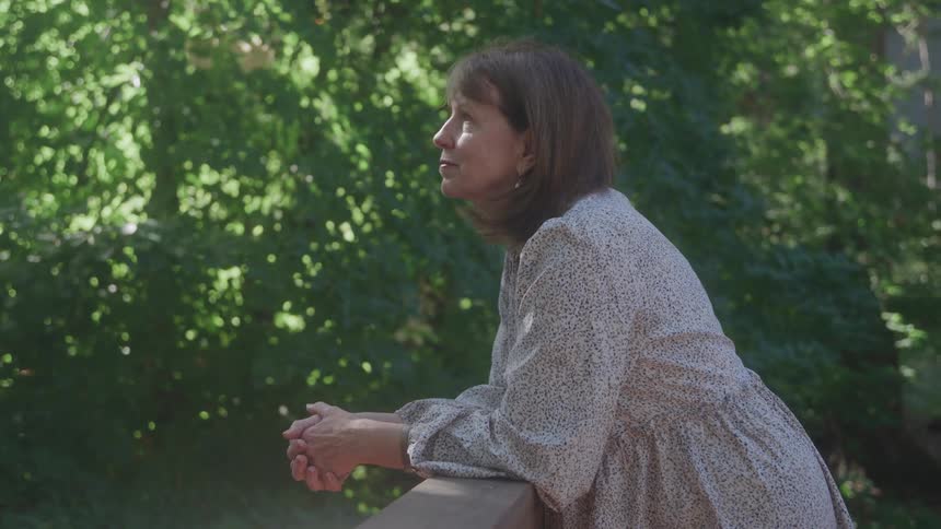 A woman in a flowing white and black patterned dress leans on a railing outdoors looking up and smiling softly with bright sunlight shining through green trees