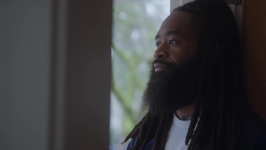 Man with a long beard and dreadlocks looks outside near the door