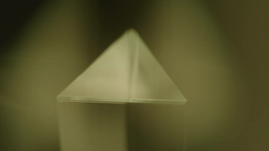 A clear threedimensional geometric shape possibly a prism or pyramid tip is looking forward and is sharply defined against a changing color background