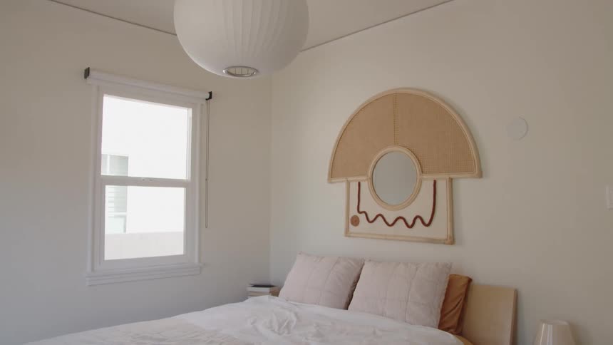 A bedroom is softly lit at midday sunlight streaming through a square window a round ceiling light hangs overhead on the bed rest two pillows neatly arranged while a mirror art design decorates the wall