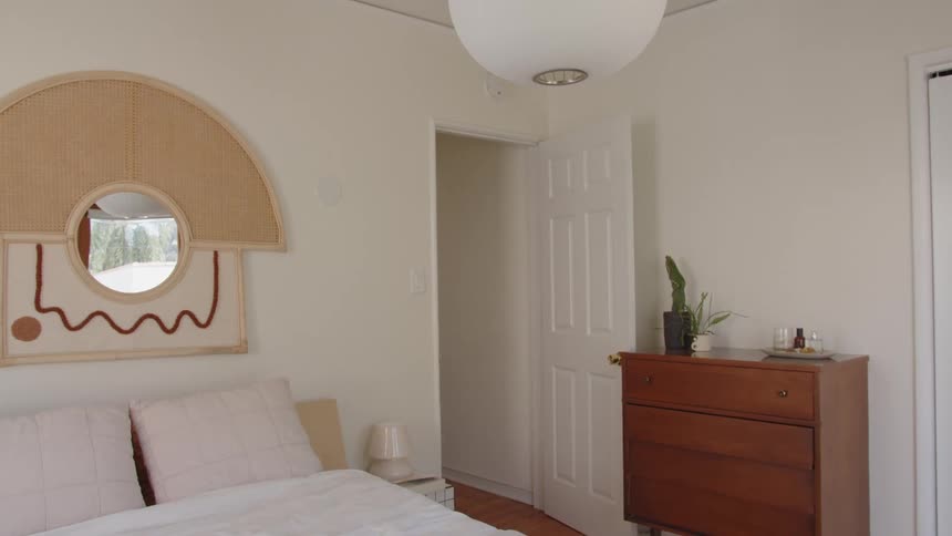 The video shows the corner of a bright minimalist bedroomon the left there is a bed with lightcolored pillows and a unique semicircular woven rattan mirrorheadboard with an abstract embroidered design hanging above iton the right there is a
