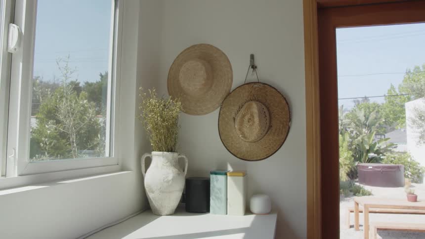 Sunlight streams into a bright airy corner of a home highlighting two straw hats hanging on the wall and a decorative vase next to a window overlooking a patio