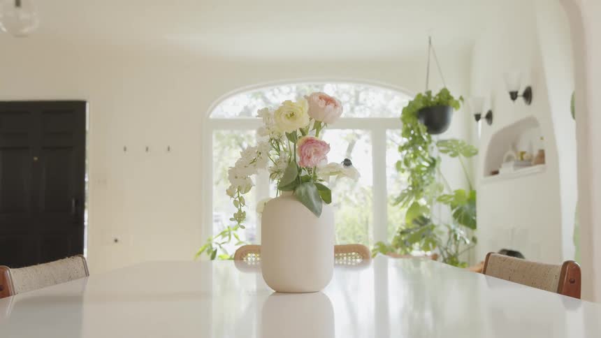A white vase containing a pink and white flower bouquet sits on a dining room table