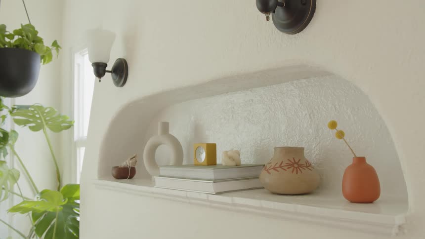 Sunlight softly illuminates an arched wall nook decorated with earthy ceramic vases books and a single yellow flower creating a calm and serene interior space