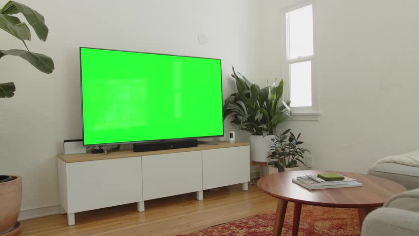 The video shows a bright modern living room with a large television screen displaying a chroma key green color suitable for digital effectsthe tv rests on a white and light wood media console below a soundbar with large potted plants flanking the s
