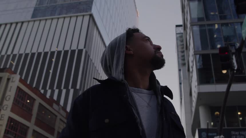A young man wearing a hoodie stands on a city street at dusk gazing upward at the towering skyscrapers with a contemplative expression