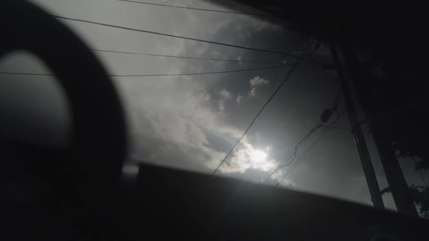 Sunlight struggles to break through dark drifting clouds behind silhouetted power lines viewed through the windshield of a stationary vehicle as a wiper rhythmically clears the glass