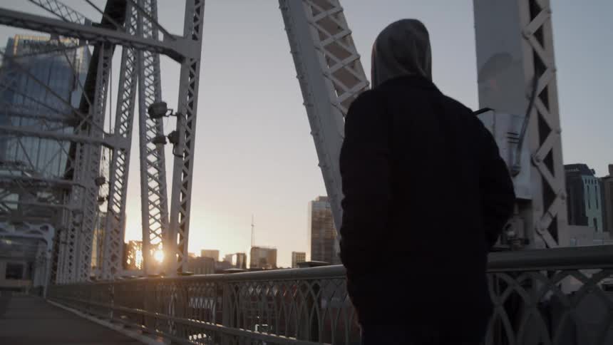 A man walks across a steel bridge toward a city skyline at sunset