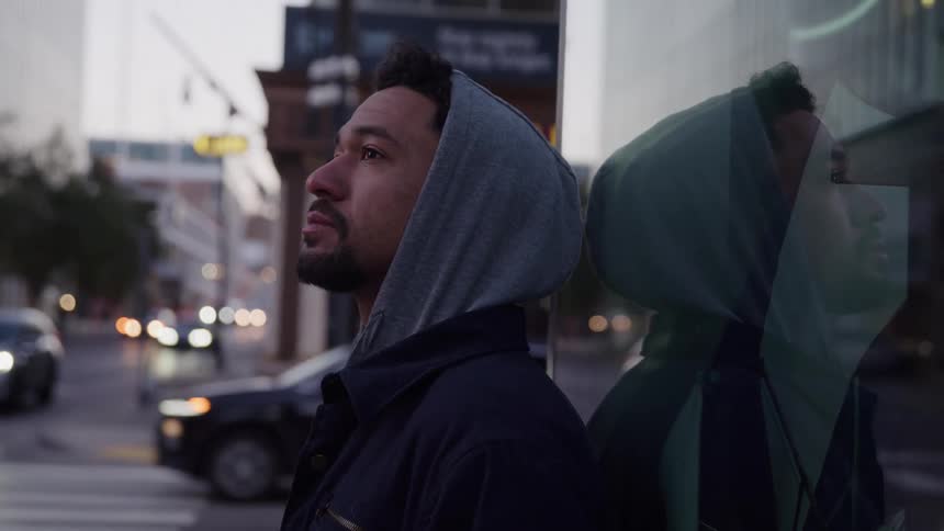 A man in a hoodie leans against a glass wall looking upward on a city street at dusk