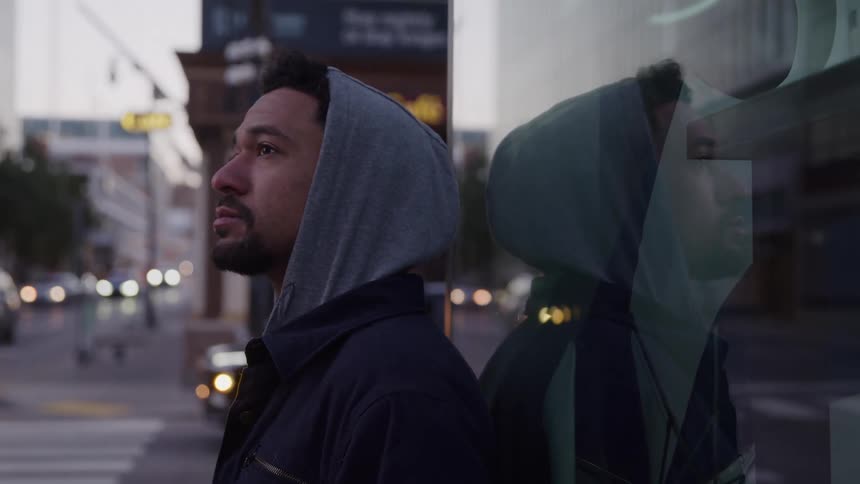 A man wearing a hooded sweatshirt leans against a window in a city at dusk looking contemplative as the streetlights and traffic reflect in the glass