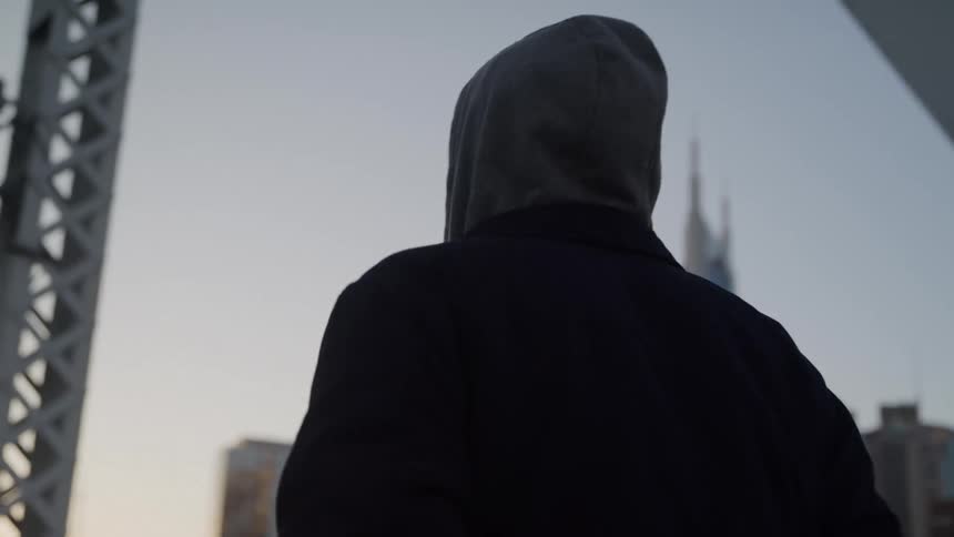 Man wearing a hooded jacket walks across a bridge with city buildings in the distance