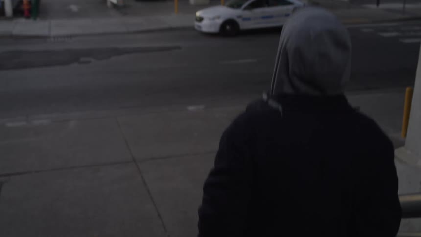 A man wearing a dark jacket and a gray hooded sweatshirt walks down a set of stairs leading away from a large concrete overpass structure in an urban setting at dusk