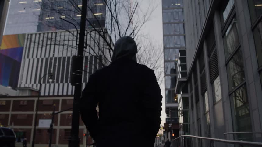 A person wearing a dark hoodie walks purposefully down a dim city sidewalk at dusk flanked by tall glass skyscrapers and bare trees that line the quiet urban street