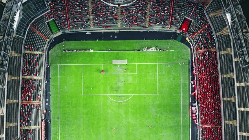 A stunning topdown aerial view slowly pulls back to reveal an entire soccer stadium filled with cheering fans as players compete on the bright green field below