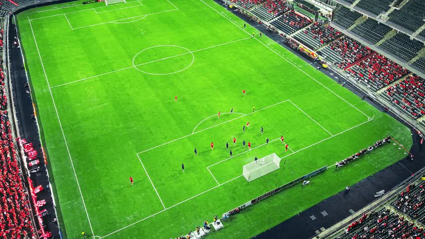 The video shows a highangle shot of a football match being played in a large stadiumplayers in red and blue uniforms are maneuvering near the penalty box and goal on the lower right side of the green fieldthe stands are partially visible and appear