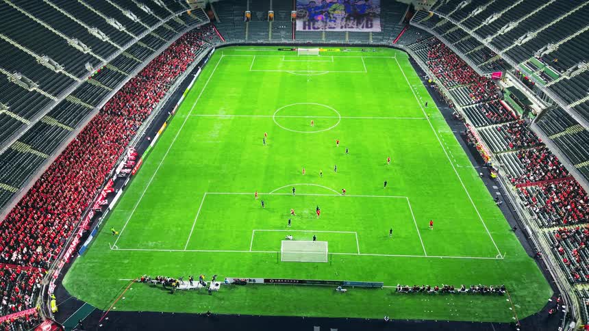 From a high vantage point a live soccer match unfolds in a vast stadium with players strategically moving across the bright green pitch as crowds cheer from the stands