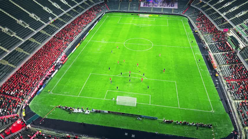 The video provides an elevated panoramic view of a soccer stadium during a matchthe bright green pitch is centered with players in red and dark blue uniforms gathered primarily near the goal on the lower half of the screenthe surrounding stadium