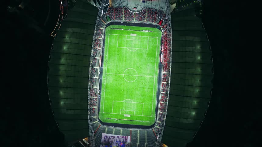 An extreme highangle aerial drone shot of a brightly lit soccer stadium at night showing the entire green field and the densely packed red and dark seating areas filled with spectators