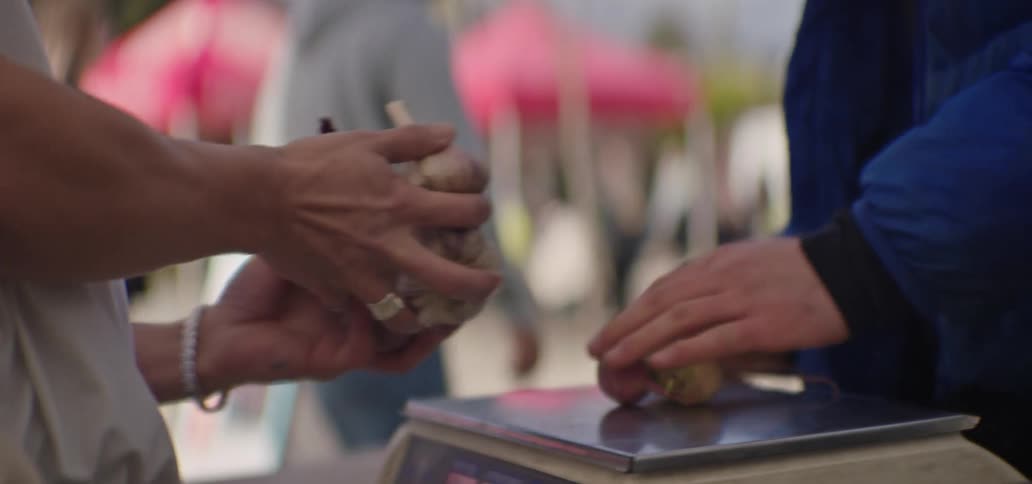 A closeup view shows a shoppers hands placing a red onion and a head of garlic onto a weighing scale held by a vendor in a blurred outdoor farmers market setting