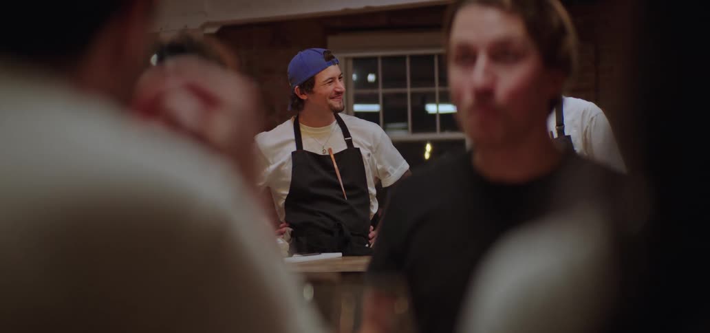 Cook wearing an apron and a blue cap stands in the background while people dine in the foreground
