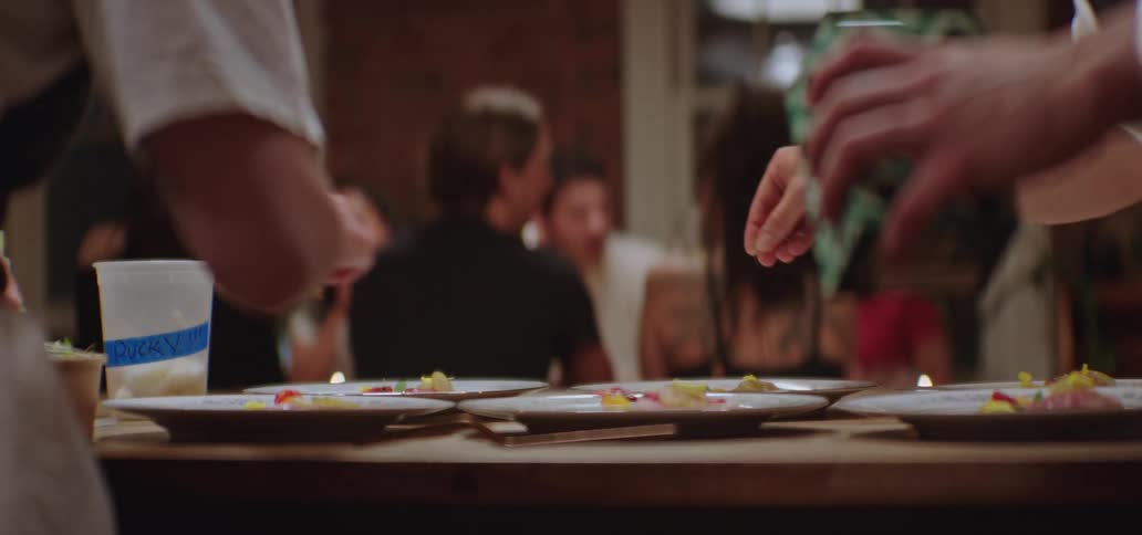 Two cooks placing items on plates of food on the counter as people sit at a table in the distance