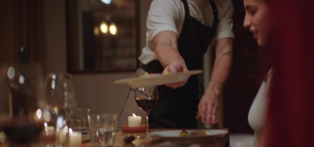 At an intimate dinner party a server sets down a plate with a colorful appetizer the warm glow of candlelight reflecting in the surrounding wine glasses