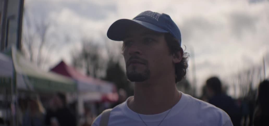 An adult male wearing a blue baseball cap and a white shirt is looking forward with interest in a crowded outdoor marketplace under a cloudy sky