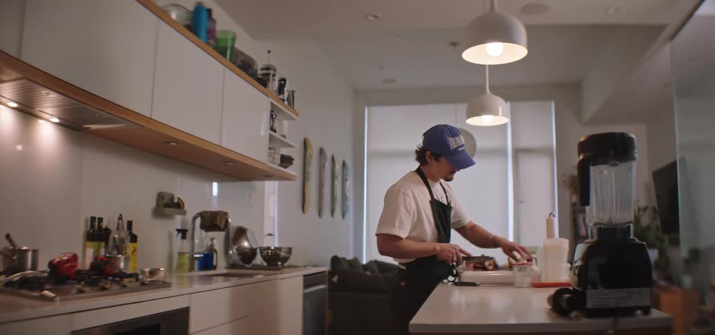 A male wearing an apron and a baseball cap is looking forward with focus while preparing food on a white counter in a modern welllit kitchen
