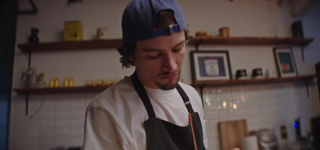 Cook wearing a blue cap uses a mandoline to slice food while another person works in the background