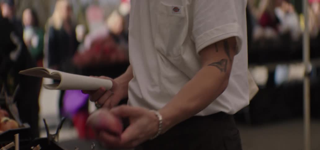 A man in a white t shirt and blue cap writes in a notepad and inspects an onion at a market
