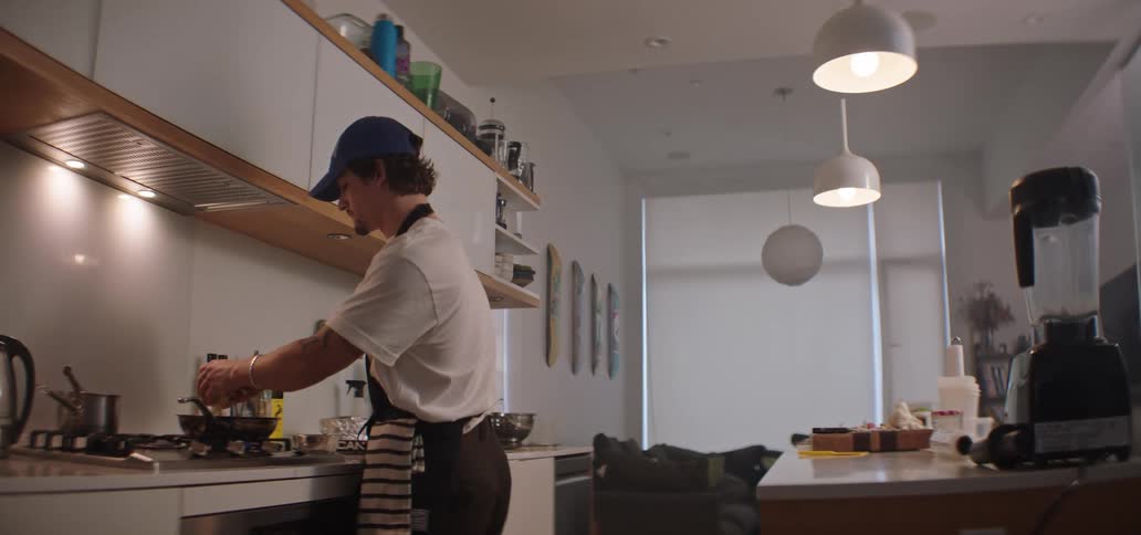 A man wearing a baseball cap and apron works in a spacious modern kitchen moving between preparing food at the island counter and cooking on the stovetop under bright overhead lighting