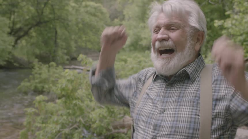 An old man laughs and claps in the forest by a river