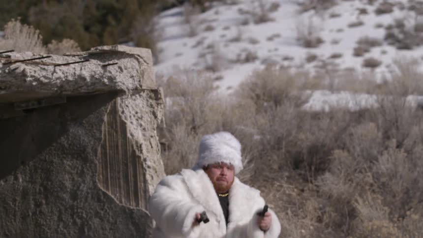 A man wearing a fur coat jumps and fires guns