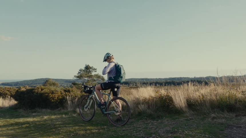 Young woman sits on her bicycle in a rural field adjusting the straps on her backpack
