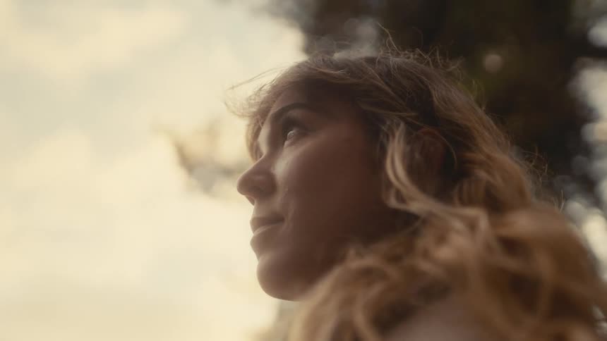 The video is a closeup lowangle shot of a young womans face profile to the left looking up towards the sky she has wavy sunkissed hair and is positioned against a bright hazy sky with dark foliage suggesting she is outdoors in a natural setting