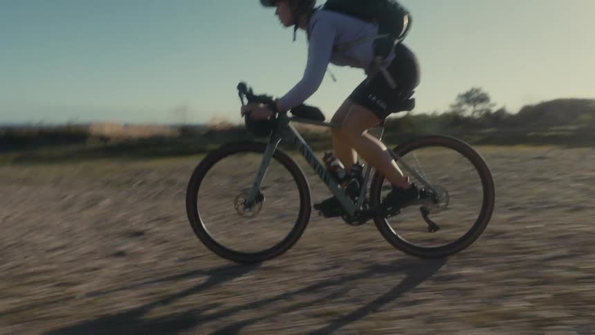 A young woman riding a bike across a field at midday wearing a helmet and carrying a backpack