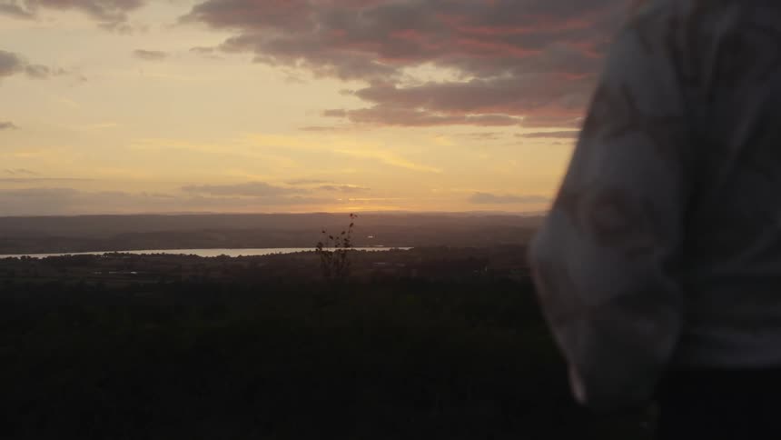 From a darkened hilltop a person gazes out at a vast valley where a river reflects the warm golden light of a beautiful sunset