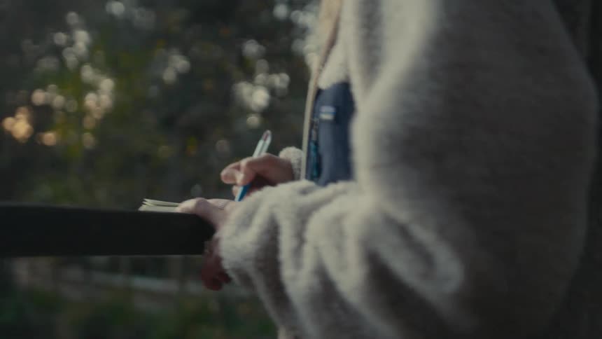 A woman standing at a railing fence writing with a pen on paper placed on the rail surrounded by trees in the woods at midday