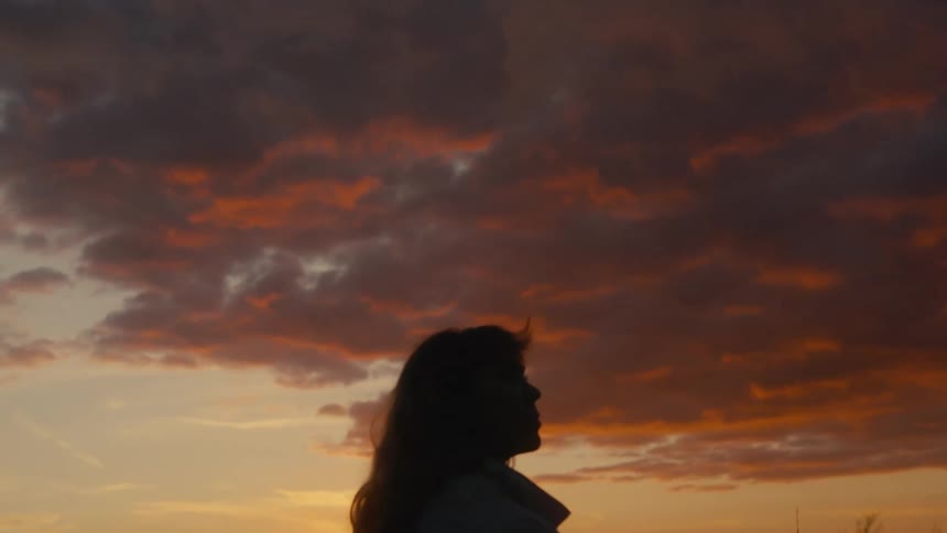 The video shows a silhouette of a person standing outside with their back mostly to the camera looking towards a dramatic sunset the sky is filled with dark textured clouds richly illuminated by orange and red hues